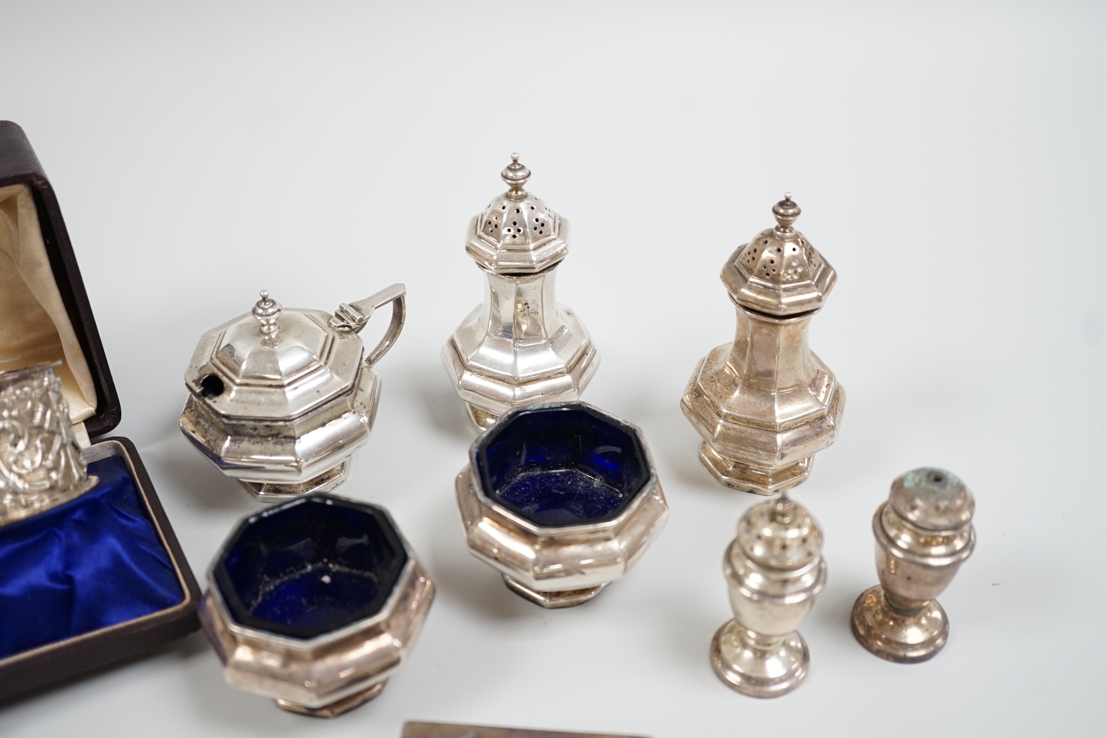 A cased pair of Edwardian embossed silver napkin rings, Birmingham, 1904, a five piece silver condiment set, Birmingham, 1926, two plated condiments and a match sleeve.